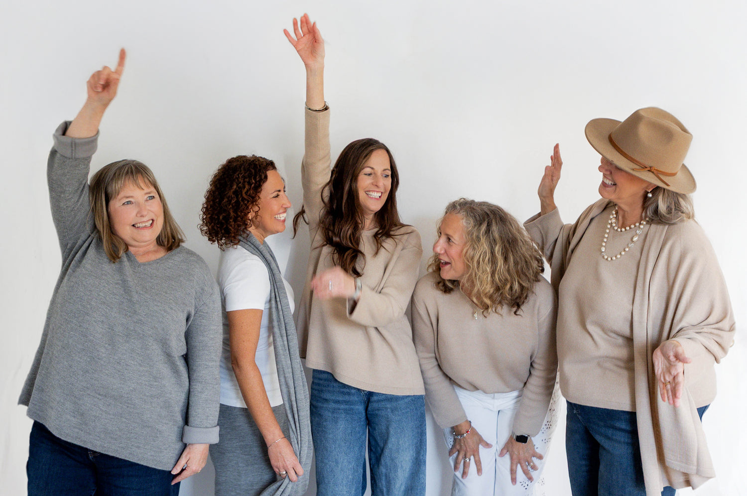 Five women standing together smiling with arms raised against a white background