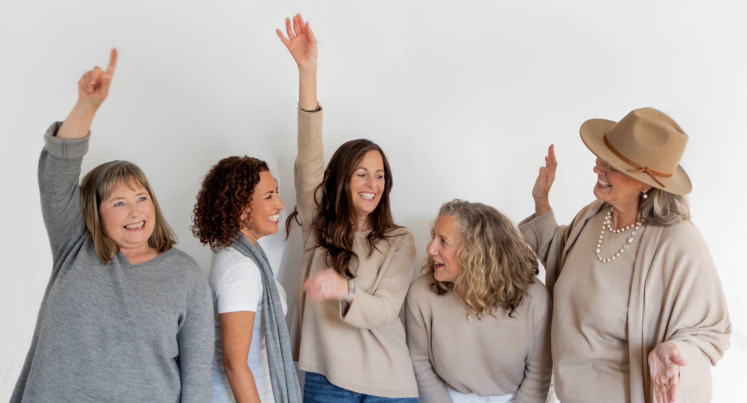 Five women standing together smiling with arms raised against a white background